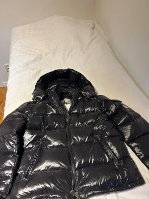 Svart glansig pufferjacka Moncler - Säljer en svart glansig pufferjacka från Moncler med huva och dragkedja framtill. Jackan har två snedställda fickor med dragkedja och en Moncler-logga på ärmen. Perfekt för kalla vinterdagar och riktigt snygg streetstil.