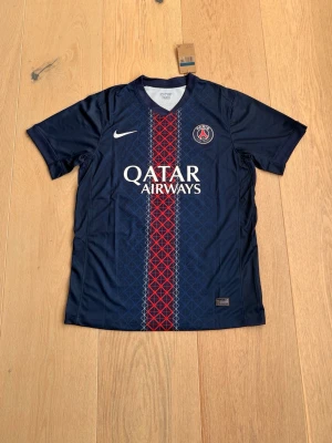 PSG matchtröja O. Dembélé #10 - Snygg mörkblå PSG matchtröja från Nike med Qatar Airways-logga på bröstet och klubbmärke. Röd och blå mönstrad rand i mitten. På ryggen står O. Dembélé och nummer 10. Perfekt för fotbollsfans och sportaktiviteter.