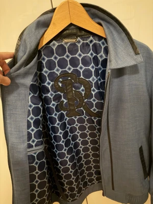 Blå jacka från Billionaire med läderdetaljer - Snygg blå jacka från Billionaire med svarta läderdetaljer vid krage och fickor. Insidan har ett unikt hexagonmönster i blått och vitt samt ett stort broderat BR-märke. Jackan har dragkedja och ribbade muddar.