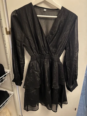 Svart glittrig omlottklänning XS - Snygg svart kort omlottklänning med glittriga detaljer och lång ärm. Klänningen har markerad midja och v-ringning, perfekt för dig som vill sticka ut. Tunt, lätt material som ger en luftig känsla.