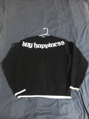 Svart stickad tröja med tryck - Cool svart stickad tröja med vit text 'buy happiness' på ryggen och en stor vit $-symbol framtill. Tröjan har rund hals, långa ärmar och vita detaljer vid ärmslut och nederkant. Perfekt för dig som gillar streetstyle och statement-plagg.