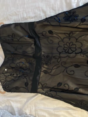 Svart blommig axelbandslös baby doll  klänning - Säljer en svart axelbandslös klänning med transparent tyg och blommigt mönster. Klänningen har en markerad midja och lätt utsvängd kjol. Perfekt för dig som vill ha en elegant look med fina detaljer.