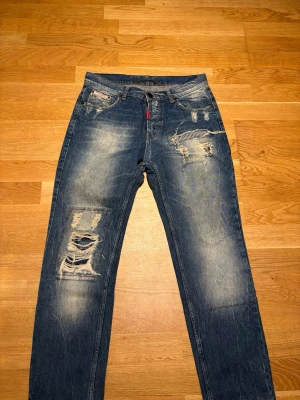 Blå jeans från Adrian Hammond - Säljer ett par blå jeans från Adrian Hammond med tydliga slitningar och hål på benen från fabrik. Jeansen har klassisk femficksmodell, dragkedja och knapp, samt en röd Hammond-tag vid gylfen. Materialet är denim  med en cool, avslappnad look. Strl 32