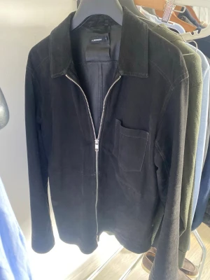 Svart overshirt från jlindeberg - Stilren svart overshirt från llindeberg i mjukt mockaimitationsmaterial. Jackan har en rak passform, klassisk krage, bröstficka och dragkedja framtill. Perfekt lager-på-lager-plagg för en clean och modern look.