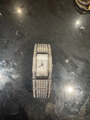 Glittrig silverklocka från D&G - Säljer en riktigt snygg och glittrig klocka från D&G i silverfärg. Klockan har rektangulär urtavla och är täckt med massor av gnistrande stenar både på armbandet och urtavlan. Perfekt accessoar för dig som vill sticka ut och glänsa.