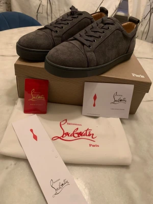 Grå sneakers från Christian Louboutin - Snygga grå sneakers från Christian Louboutin med klassisk låg profil och ikonisk röd sula. Skorna är tillverkade i mocka med matchande grå skosnören och detaljer. Perfekt för dig som vill ha en exklusiv och stilren look.
