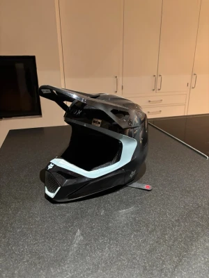 Svart hjälm från Fox  - Säljer en svart hjälm från Fox med ljusblått logotryck och detaljer. Hjälmen har ett robust visir framtill och ventilationshål för bättre komfort. 