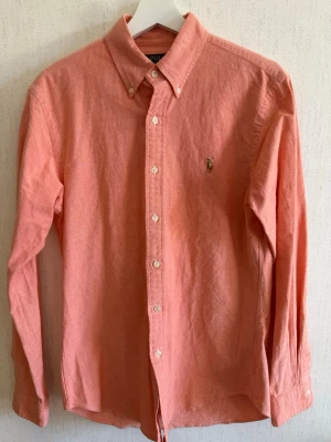 Korallrosa skjorta från Polo Ralph Lauren - Snygg korallrosa skjorta från Polo Ralph Lauren i slim fit-modell. Skjortan är i bomull med stretch, har klassisk krage, knappar framtill och broderad logga på bröstet. Perfekt för en stilren look.