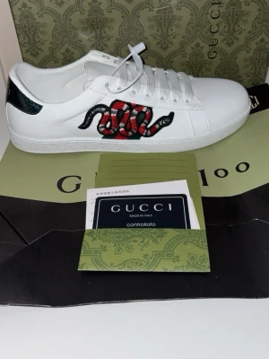Gucci ace vita snake strl 42 - Snygga vita sneakers från Gucci i läder med den ikoniska broderade ormen i rött, svart och vitt på sidan. Baksidan har gröna och röda detaljer samt svart ormskinnsmönster. Klassisk låg modell med vita skosnören och gummisula.