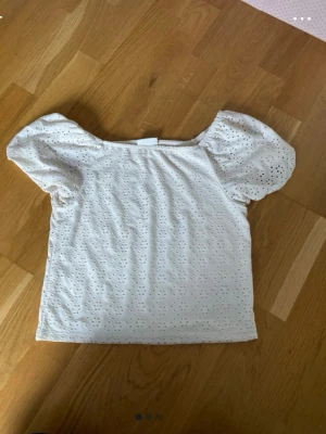 Vit blus med puffärm och broderi - Söt vit blus med korta puffärmar och broderade hålmönster över hela tyget. Toppen har en rak passform och är perfekt för en somrig look. Tillverkad i bomull för en luftig och bekväm känsla.