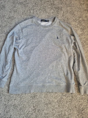 Grå sweatshirt från Polo Ralph Lauren - Klassisk grå sweatshirt från Polo Ralph Lauren med rund halsringning och broderad logga på bröstet. Tröjan har ribbade muddar vid ärmslut och nederkant. Perfekt för en avslappnad och stilren look. Passar som M.