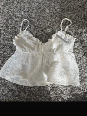 Vit linnetopp med knyt och volang - Söt vit linnetopp med tunna axelband, volangdetaljer vid bysten och knyt framtill. Toppen har en luftig och lätt struktur, perfekt för varma dagar. Passar till både jeans och kjol för en trendig look.