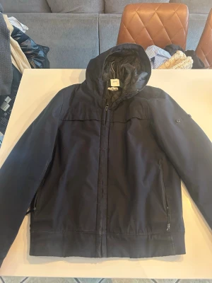 Svart vindjacka med huva från Wesc - Snygg svart vindjacka från Stone Island  med huva och dragkedja framtill. Jackan har två fickor med dragkedja och detaljer med knappar på ärmen där det saknas en badge.  Perfekt för blåsiga dagar och enkel att matcha med olika outfits.