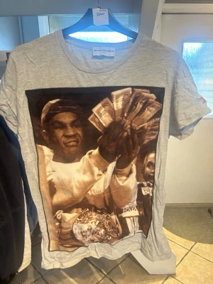 Grå t-shirt med tryck och palmlogga - Grå t-shirt från Bastille  Rive Droite med stort tryck framtill som föreställer en person med sedlar och bälte. Liten röd stjärna med svart palm på ärmen. Rund halsringning och avslappnad passform.