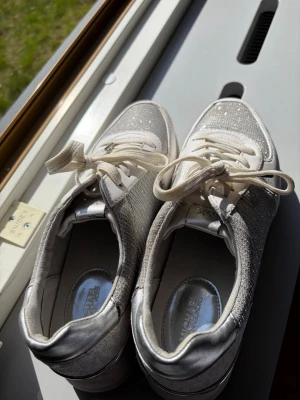 Glittriga sneakers från Michael Kors - Snygga sneakers från Michael Kors i silver och vitt med glittriga detaljer. Skorna har klassisk låg modell, vita snören och metallic-finish på häl och sidor. Perfekta för dig som vill sticka ut med stilrena och trendiga skor.