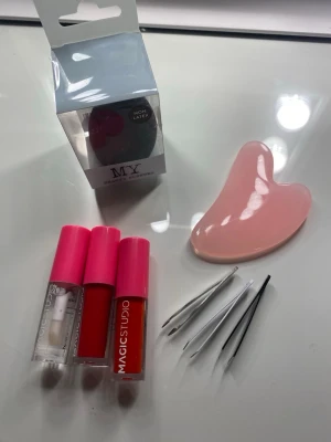Skönhetstillbehör mix med gua sha - Ett kit med skönhetstillbehör: rosa gua sha-sten, tre pincetter i metall, tre läpprodukter från Magic Studio med rosa lock samt en makeupsvamp från MY i originalförpackning. Perfekt för ansiktsvård och sminkapplikation.