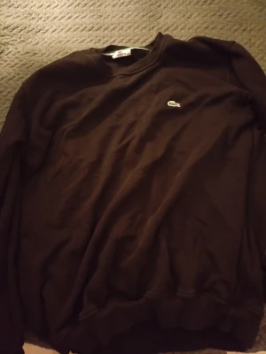 Svart stickad tröja från Lacoste - Säljer en svart stickad tröja från Lacoste med klassisk rund hals och det ikoniska gröna krokodilmärket broderat på bröstet. Tröjan har långa ärmar och är tillverkad i mjukt material som passar perfekt för en avslappnad och stilren look.