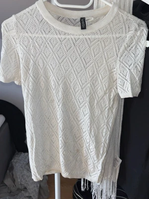 Vit spetsmönstrad topp från H&M Divided - Säljer en vit, kortärmad topp från H&M Divided med spetsmönster i geometriskt hålmönster. Toppen har rund halsringning och är gjord i ett tunt, luftigt material som passar perfekt till sommaren.