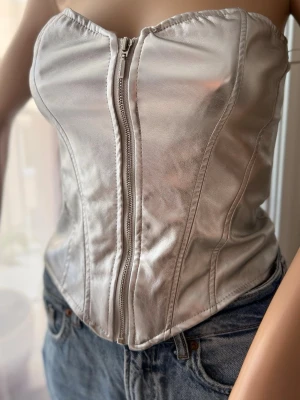Silverfärgad korsett med dragkedja - Snygg korsett i metallic silver med markerade sömmar och dragkedja framtill. Axelbandslös modell med hjärtformad urringning och figurnära passform. Perfekt att styla till jeans för en cool look.