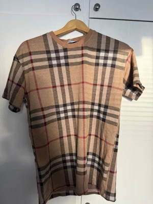 Burberry Set - Snyggt tvådelat set med t-shirt och shorts i beige med klassiskt rutigt mönster i svart, vitt och rött. T-shirten har rund hals och korta ärmar, shortsen har resår i midjan och sidofickor. Perfekt för en avslappnad och trendig look.