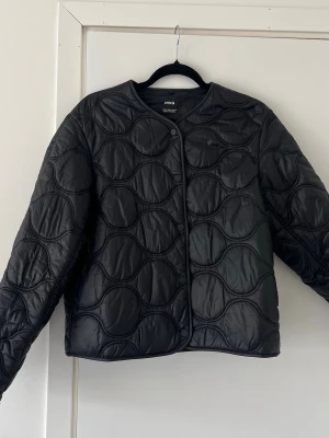 Svart quiltad pufferjacka från MNG med fickor - Snygg svart pufferjacka från MNG med vågformat quiltat mönster. Jackan har rund halsringning, tryckknappar framtill och en boxig passform. Tillverkad i glansigt syntetmaterial som ger en modern look. Perfekt till våren då den inte är supervarm. 🥰