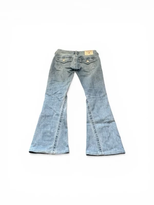Ljusblå bootcut jeans från True Religion - Säljer ett par Lågmidjade ljusblå bootcut jeans från True Religion med klassiska fickor och snygga detaljer bak. Jeansen har utsvängda ben och en cool tvätt framtill. Perfekta för dig som gillar retrostil och vill sticka ut. Har en liten lagning men syns knappt. Har inget måttband så kan ej ge mått.