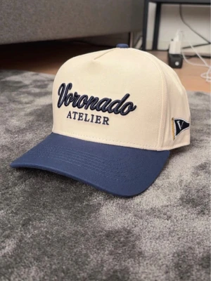 Beige och marinblå keps Voronado - Snygg keps från Voronado Atelier i beige med marinblå skärm. Broderad logga framtill och liten patch på sidan. Klassisk form med böjd skärm och justerbar baksida. Perfekt accessoar för en cool och avslappnad stil.