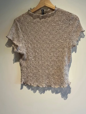 Beige spetstopp - Säljer en beige spetstopp från H&M med blommigt mönster och korta ärmar. Toppen har en lätt vågig kant vid hals, ärmslut och nederkant samt en figurnära passform. 
