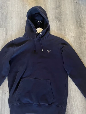Marinblå hoodie från Gant - Klassisk marinblå hoodie från Gant med broderad logga på bröstet. Den är använd ett fåtal gånger pga för liten. Storlek S
