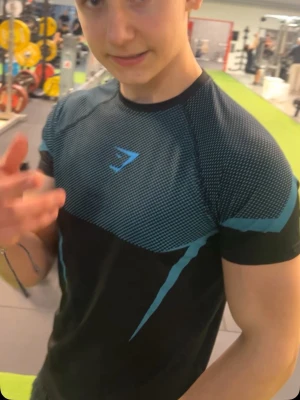 Gymshark Onyx - Snygg svart träningströja från Gymshark med blått mönster och logga på bröstet. Tröjan har korta ärmar och är tillverkad i ett stretchigt funktionsmaterial som andas, perfekt för gym och träning. Unik design med blå detaljer på ryggen. Använd en gång