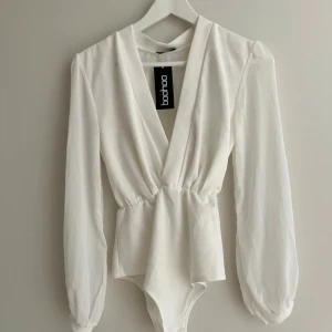 Vit omlott bodysuit från Boohoo - Säljer en vit omlott bodysuit från Boohoo med långa, luftiga ärmar i chiffong. Djup v-ringning och markerad midja ger en snygg siluett. Perfekt att styla med jeans eller kjol. Helt oanvänd                                                                      (Läs alltid min profil för villkor)