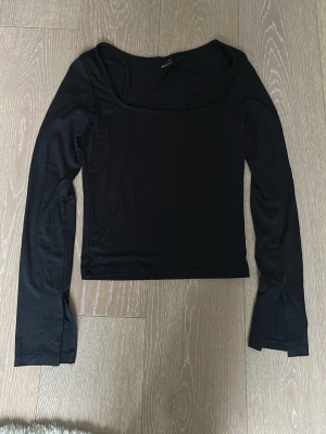 Svart långärmad topp med vid urringning - Säljer en svart långärmad tröja med vid, rund urringning. Modellen är figurnära och har långa ärmar med lätt utsvängda ärmslut. Perfekt att styla med jeans eller kjol för en clean look.