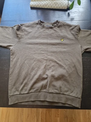 Beige sweatshirt från Lyle & Scott - Snygg beige sweatshirt från Lyle & Scott med klassisk rund hals och ribbade muddar. Tröjan har det ikoniska gula fågel-loggan broderad på bröstet. Perfekt för en avslappnad och stilren look.