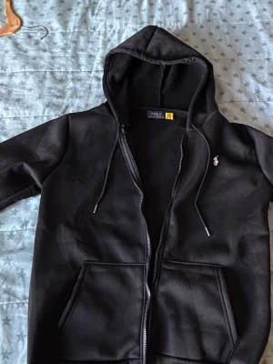 Svart hoodie från Polo Ralph Lauren - Svart hoodie med dragkedja från Polo Ralph Lauren. Klassisk design med vit broderad logga på bröstet, huva med dragsko och två stora fickor framtill. Perfekt för en chill och stilren look.