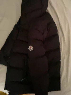 Svart dunjacka från Moncler med huva - Säljer en svart dunjacka från Moncler med huva och klassisk Moncler-logga på ärmen. Jackan har breda, vadderade paneler och dragkedja framtill. Perfekt för kalla vinterdagar och riktigt snygg streetstil.storlek 11-12y = kan passa xs eller mindre s