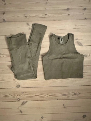 Olivgrönt träningsset STRONGER - Olivgrönt träningsset från STRONGER med ribbad struktur. Setet innehåller ett sportigt linne med breda axelband och matchande höga tights. Materialet är stretchigt och sömlöst för extra komfort under träning. Diskret logga på linning och linne.