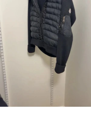 Svart pufferjacka från Moncler - Svart pufferjacka från Moncler med stickade ärmar och quiltad front. Jackan har dragkedja framtill, en ficka med dragkedja på ärmen och Moncler-logga på axeln. Perfekt för kyliga dagar med sin kombination av värmande material och snygg design.