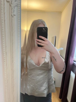 Champagnefärgad satinlinnetopp - Säljer en champagnefärgad linnetopp i satin med smala axelband och v-ringning. Toppen har en snygg knytning i ryggen och lätt glansigt material som ger en lyxig känsla. Perfekt att styla med jeans eller kjol för en trendig look. 