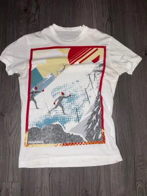 Vit Dsquared2 t-shirt med skidmotiv - Vit t-shirt från Dsquared2 med färgstarkt tryck framtill som visar skidåkare i backen och grafiska detaljer i rött, blått och gult. Klassisk passform med rund hals och korta ärmar. Perfekt för dig som gillar statement-plagg och coola prints.