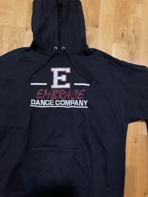 Hanes Svart Embrace Dance Company hoodie - Svart hoodie med stor huva och dragsko, samt tryck framtill med texten 'E Embrace Dance Company' i vitt och vinrött. Klassisk känguruficka framtill och ribbade muddar. Perfekt för dig som gillar streetstyle och dans.