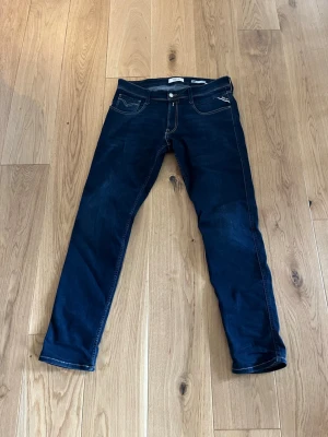 Mörkblå slim fit jeans från Replay - Snygga mörkblå slim fit jeans från Replay i modell Anbass med klassiska fem fickor och kontrastsömmar. Jeansen har en modern passform och är tillverkade i stretchigt denim för extra komfort. Perfekta för dig som gillar en stilren och trendig look.