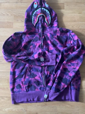 Lila camo hoodie från BAPE - Säljer en lila camo-hoodie från BAPE med hajmotiv på huvan och broderade bokstäver 'WGM' på sidan. Hoodien har dragkedja, fickor och detaljer i svart, rosa och lila. Perfekt för dig som gillar streetwear och vill sticka ut.