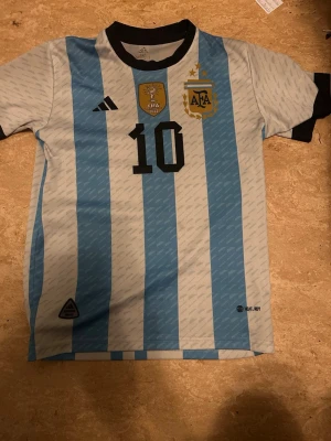 Argentina Messi matchtröja Adidas - Säljer en officiell Argentina fotbollströja från Adidas med Messis namn och nummer 10. Tröjan har klassiska blå och vita ränder, svarta detaljer vid krage och ärmslut samt guldiga emblem på bröstet. Perfekt för dig som älskar fotboll och Messi.