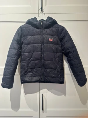 Mörkblå pufferjacka från Gant - Snygg mörkblå pufferjacka från Gant med huva och dragkedja framtill. Jackan har ett broderat Gant-märke på bröstet och två sidofickor. Perfekt för kalla dagar tack vare det vadderade materialet och den klassiska quiltade designen. Lite skada vid ena fickan, annars i super bra skick 