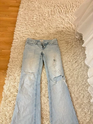 Ljusblå bootcut jeans med slitningar - Ljusblå baggy jeans med slitna detaljer och hål på båda benen. Klassisk femficksdesign och låg midja. Dom är i storlek S 14+! 