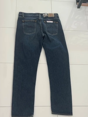 Nudie Jeans Steady Eddie - Klassiska mörkblå jeans från Nudie med rak passform och fem fickor. Jeansen har en tidlös look med kontrastsömmar och Nudie ikoniska läderpatch bak i midjan. Perfekta för en avslappnad och stilren outfit. Lapp fortfarande på och helt oanvända. Storlek w33 l32