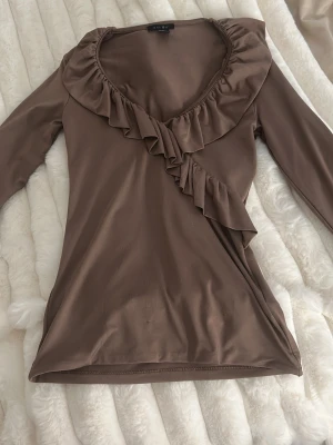 Brun volangtopp med v-ringning new yorker  - Snygg brun topp från new yorker med lång ärm och djup v-ringning. Toppen har volangdetaljer längs ringningen och framtill, vilket ger en trendig och feminin look. Perfekt att styla med jeans eller kjol för en chic outfit.