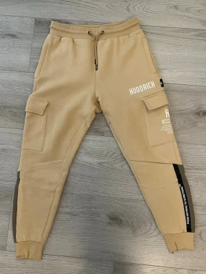 Beige cargobyxor från Hoodrich - Stilrena beige cargobyxor från Hoodrich med stora fickor på sidorna, resår och dragsko i midjan samt muddar vid bensluten. Snygga svarta detaljer längs benen och tryckt logga på ena benet. 