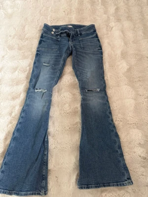 Blå bootcut jeans från Gina Tricot - Säljer ett par blå bootcut jeans från Gina Tricot i storlek XS. Jeansen har slitna detaljer på knäna och framsidan, två knappar i midjan och klassiska fickor. Perfekta för en trendig och avslappnad look.