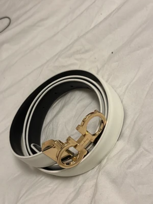 Vitt Ferragamo bälte med gulddetalj - Snyggt vitt bälte från Ferragamo med ikoniskt guldspänne. Bältet har en stilren design med svart insida och exklusiv känsla. Perfekt accessoar för att lyfta din outfit med en lyxig touch. Helt ny!